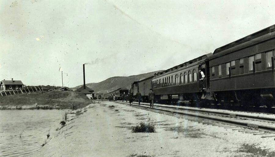 A passenger train rides along the river with the railroad station and buildings in the background. A few men walk along side the cars.