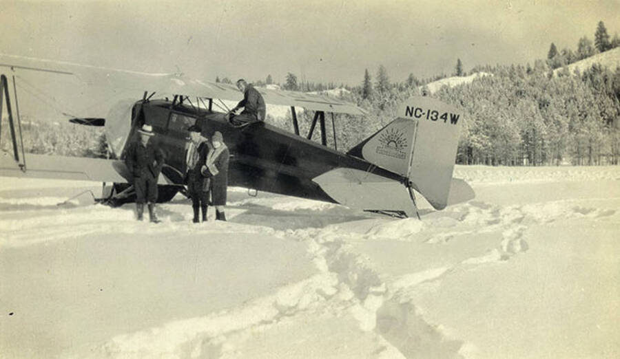 Golda Stonebraker, Al Stonebraker (middle), pilot A. A. Bennett (on plane), and an unidentified man stand in the snow beside a seven passenger Zenith airplane at Stonebraker Ranch in the Chamberlain Basin. The photo caption reads: 'Taking Al out.'