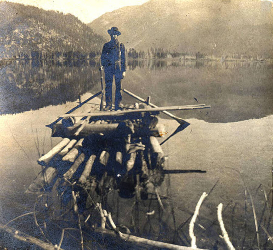 Man with rifle stands on a raft on a like with a mirror reflection.