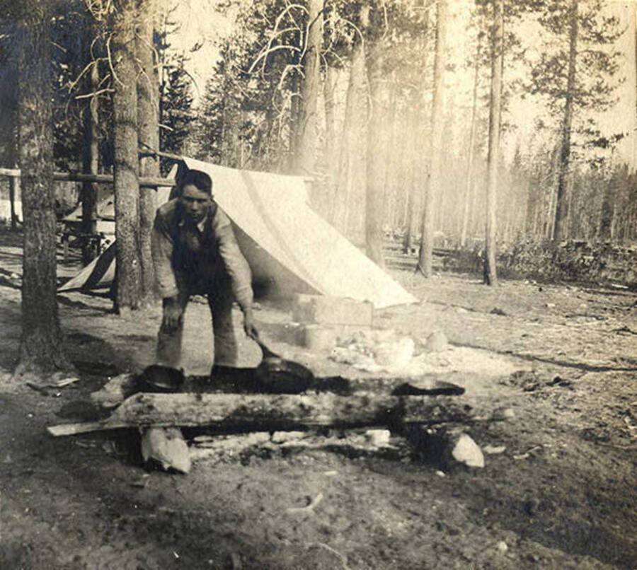 Sumner Stonebraker bent over fire hold a frying pan cooking breakfast near a tent in the woods.