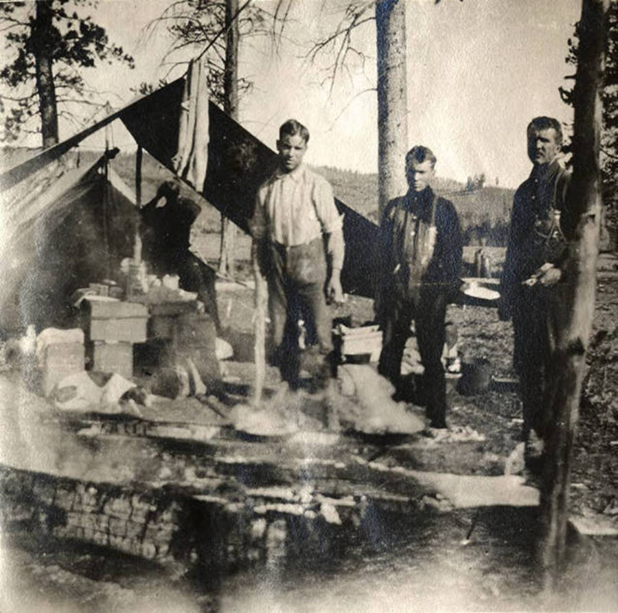 Three men stand outside a canvas tent with cooking supplies nearby.