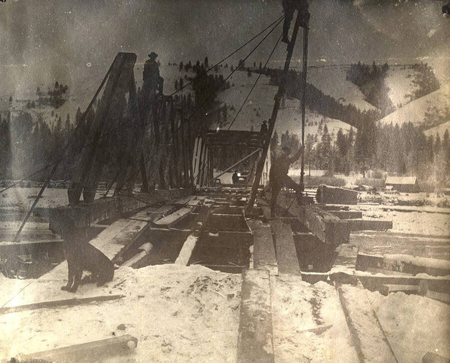 Dog sits on the snowy bridge while men work in the background.