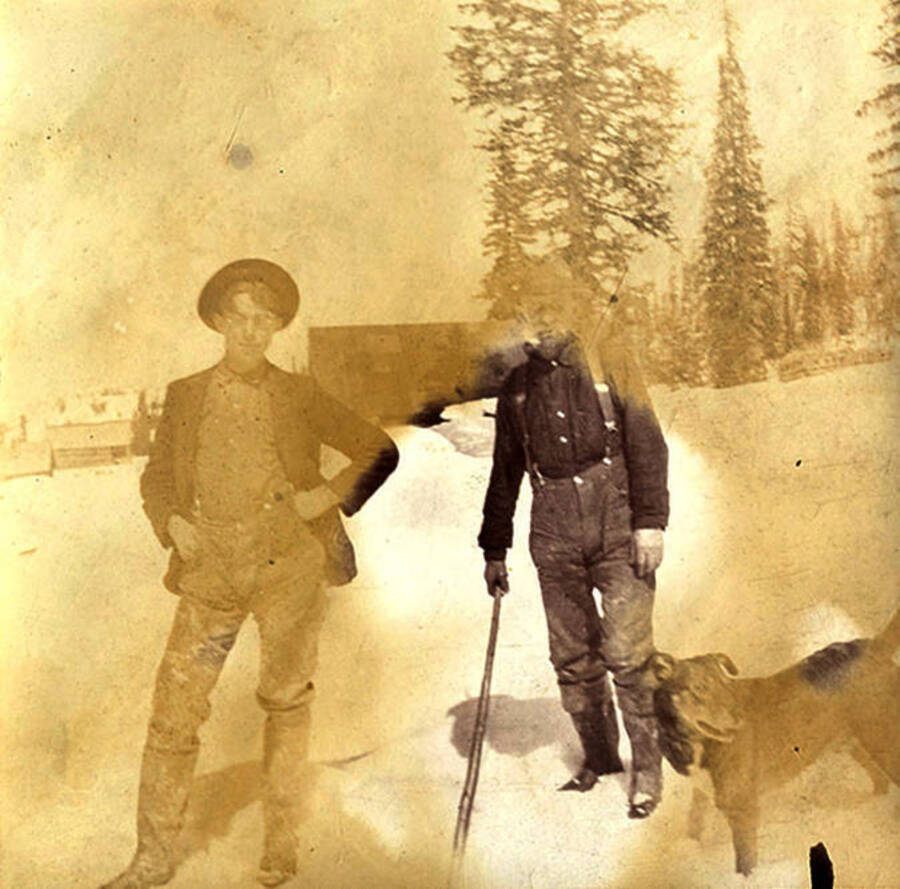 A young man and an elderly man stand in the snow with a dog. Log cabins can be seen in the background. Near Roosevelt, Idaho.