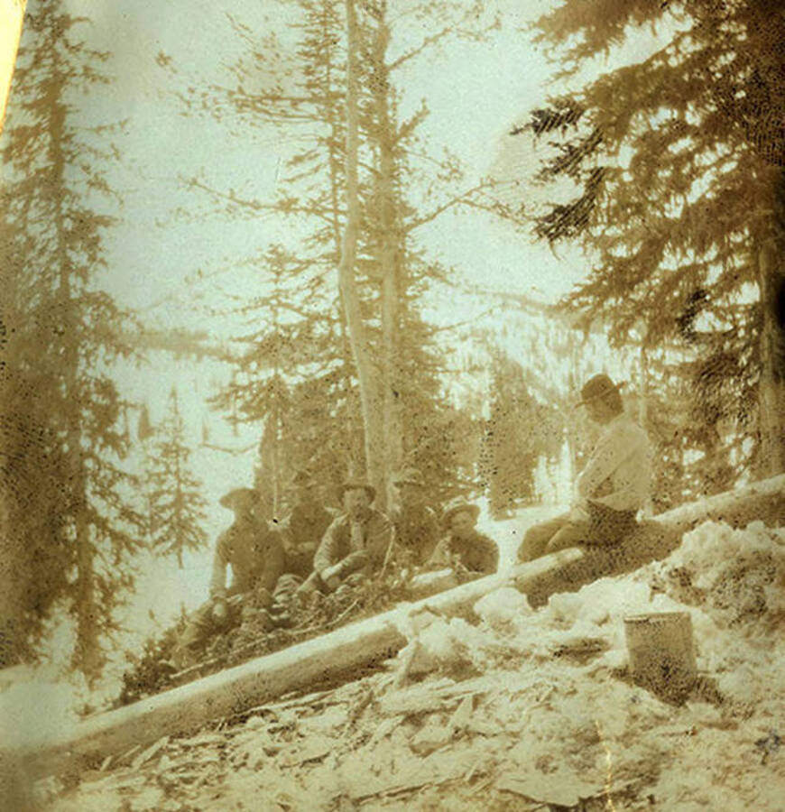 Six men wearing hats sit on logs in a snowy forested area.