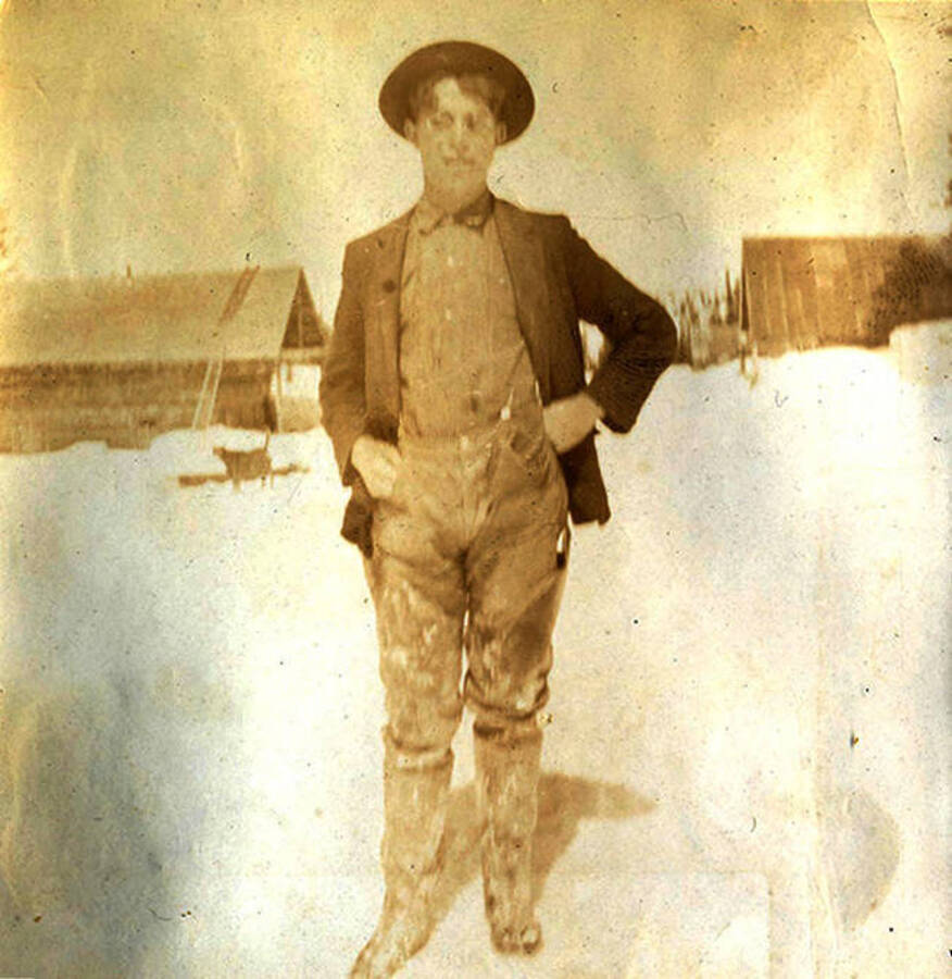Young man wearing glasses stands in the snow with log cabins in the background. Near Roosevelt, Idaho.