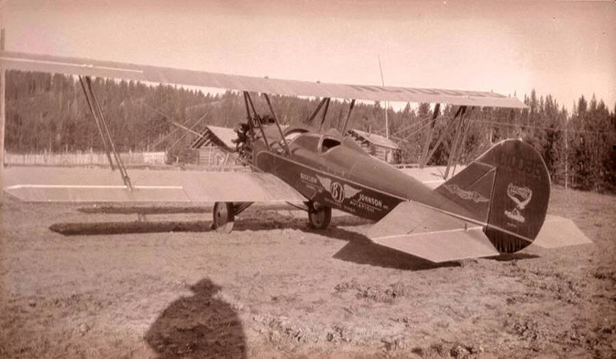 Airplane with the words 'Bigelow-Johnson School of Aviation, Spokane, Washington' sits in a meadow near the Stonebraker Ranch in the Chamberlain Basin. The homestead and corral can be seen in the background.