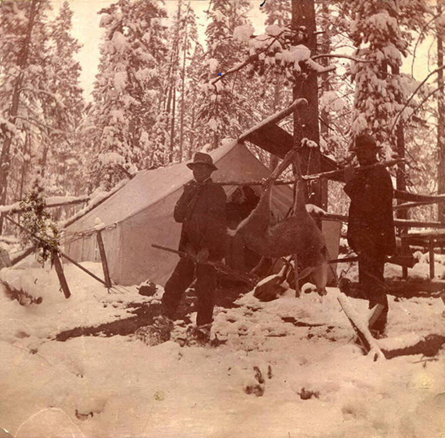 Two men pose with a harvester deer outside a canvas tent in the snow.