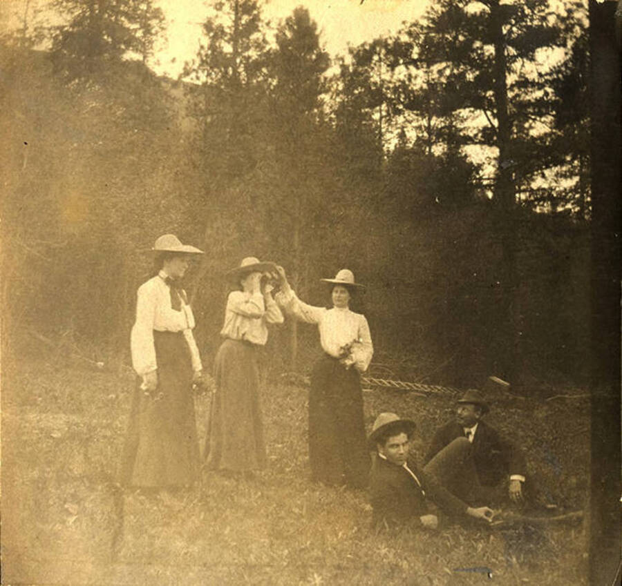 Three women and two men watch for a train using binoculars near the forest. The photo caption reads: 'Band of hoboes.'