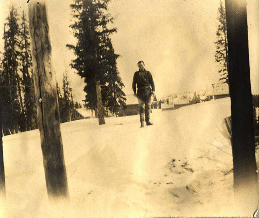 A man stands in the snow with wooden buildings in the background. Near Roosevelt, Idaho.