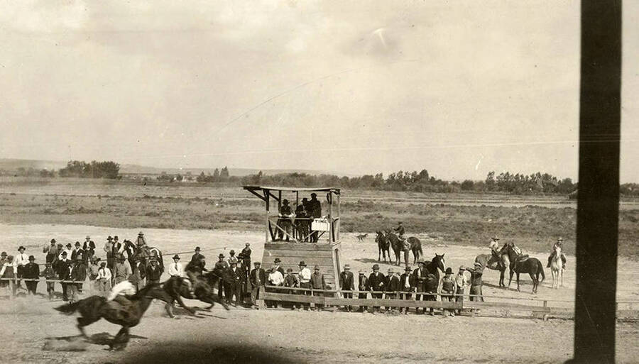 Two horses race to the finish line. Men in suits and overalls stand along the sidelines near the judges stand.