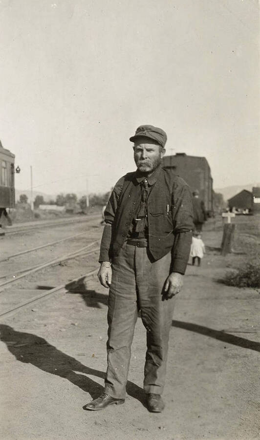 Male train conductor stands by the train tracks with a passenger car in the background.