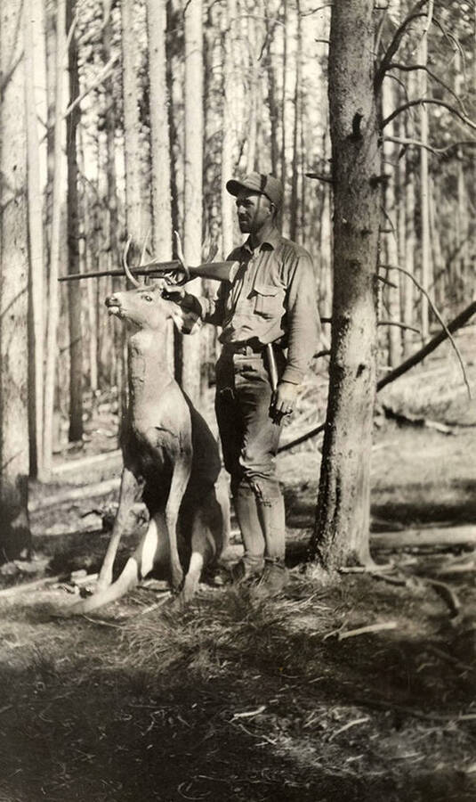 A hunter stands with his rifle beside a harvested deer in a wooded area.