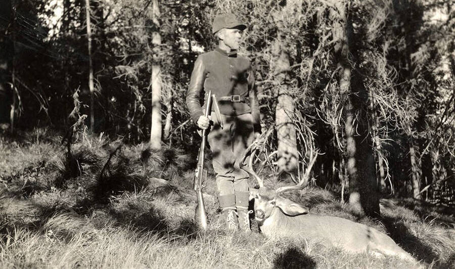 A man holds a rifle and poses with a harvested mule deer buck. 