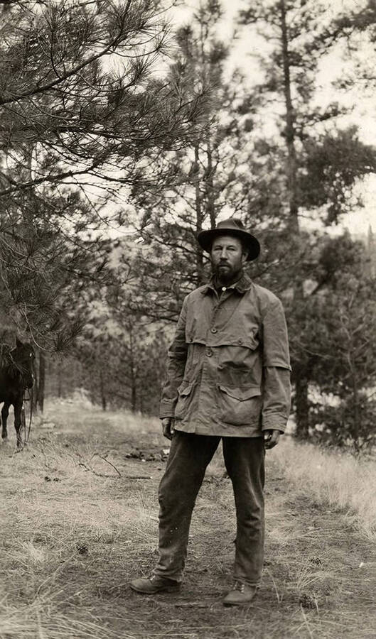 A man dressed in a jacket and a hat poses near a horse in a forested area.