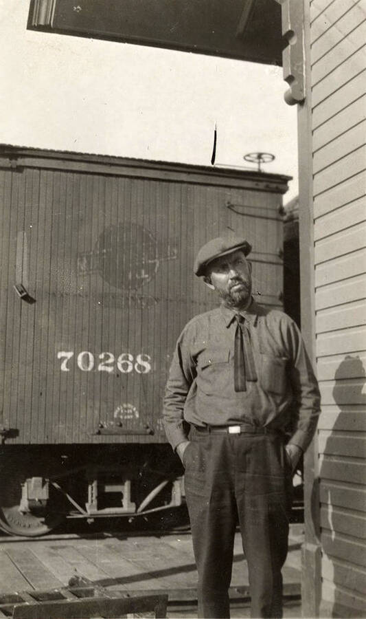 A man in a tie and a newsboy cap stands at the train station with a passenger car in the background.