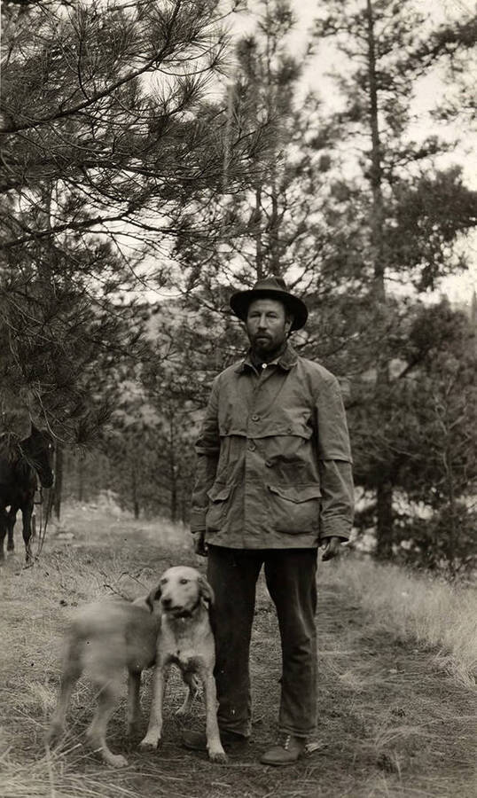 A man poses with a dog in a forested area. A horse can be seen in the background.