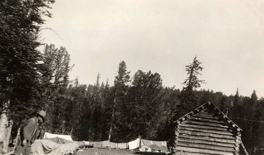 A man dressed in a hat walks by a clothesline filled with laundry in front of a log cabin.