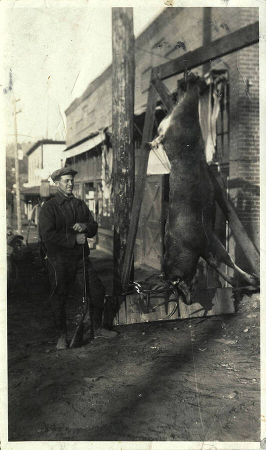 A man with a rifle stands besides a mule deer buck, hung from a wooden beam. A brick building storefront is in the background.