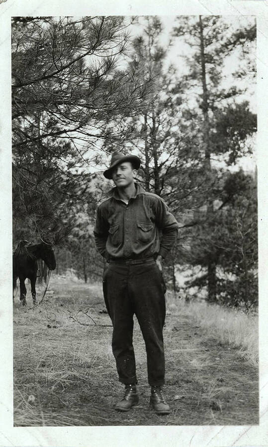 A man dressed in boots and a hat poses near a horse in a forested area.