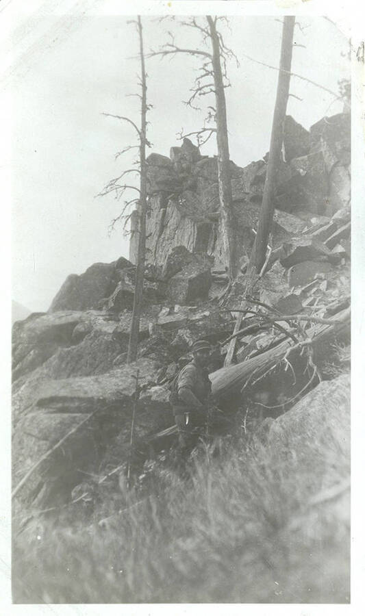 A hunter wearing a hat and carrying a rifle hikes upward through rocky terrain.