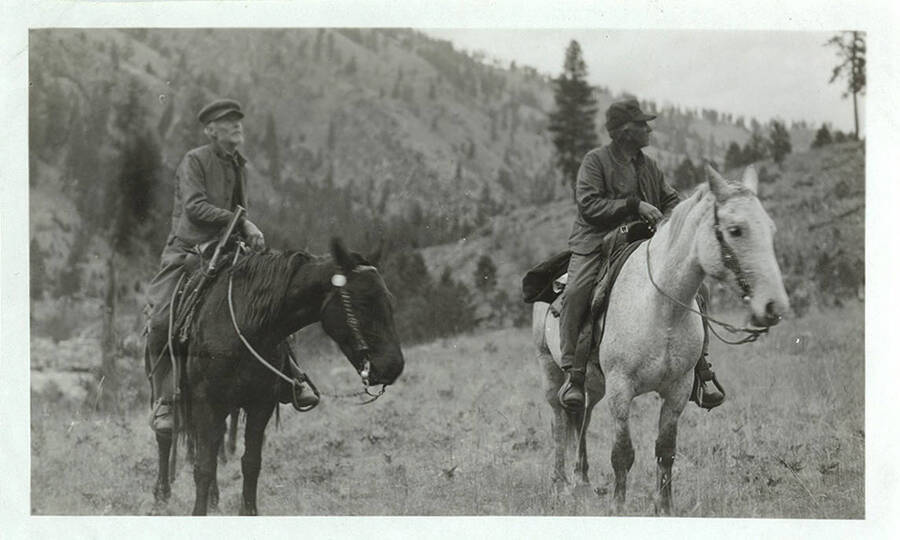 Two men ride horses and look around off into the distance.