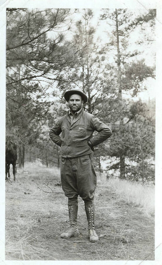 A man dressed in lace-up boots and a hat poses in a forested area. A horse can be seen in the background.