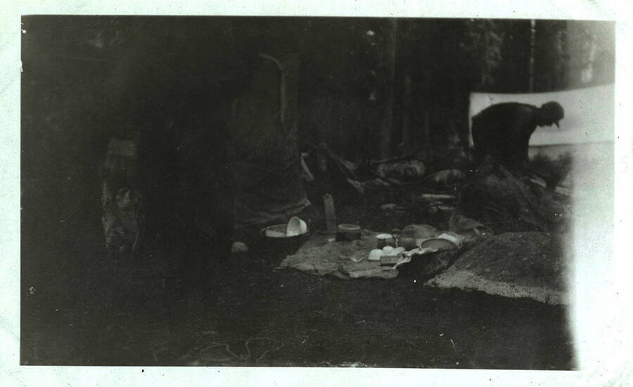 A man stands over cooking supplies and dishes near a tent at a camp site.