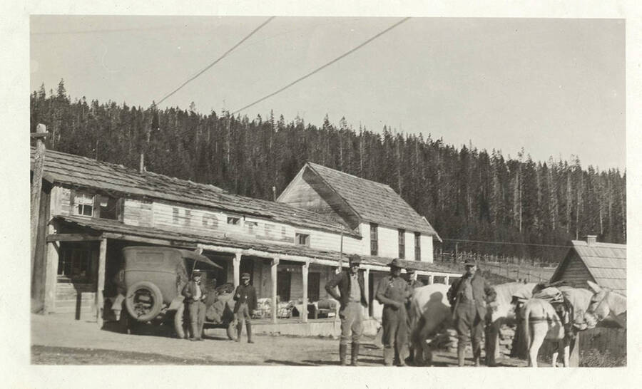 White wooden building with the word 'Hotel' printed on it located at Burgdorf Hot Springs. An automobile, six men, and a pack train stand nearby.
