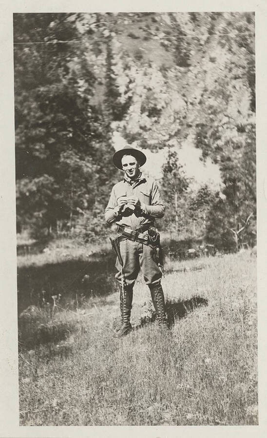 A man stands in a meadow wearing lace-up boots, a hat, and has two pistol holders around his waist.