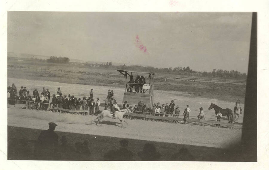 One horse crosses the finish line. Men in suits and overalls stand along the sidelines near the judges stand. Spectators in the grandstands are seen on the opposite side of the race track.