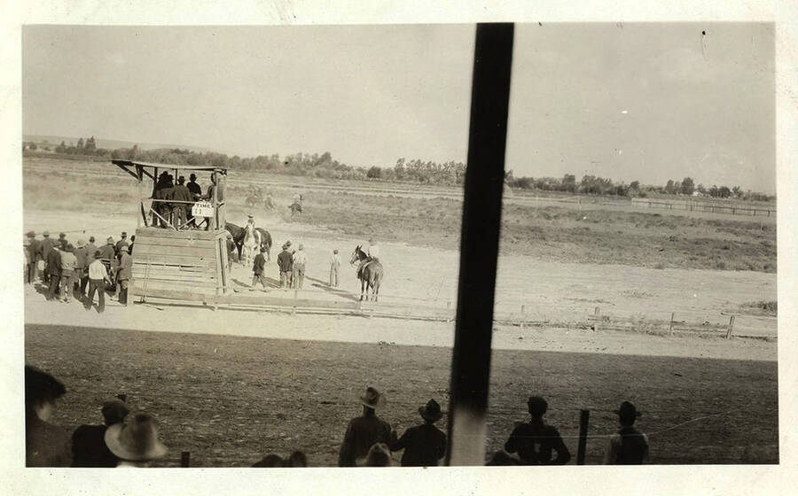 A view of a horse race track and judges station. Men in suits and overalls stand along the sidelines near the judges stand. Spectators in the grandstands are seen on the opposite side of the race track.