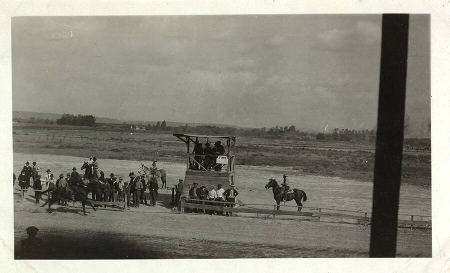 One horse crosses the finish line. Men in suits and overalls stand along the sidelines near the judges stand. Spectators in the grandstands are seen on the opposite side of the race track. 
