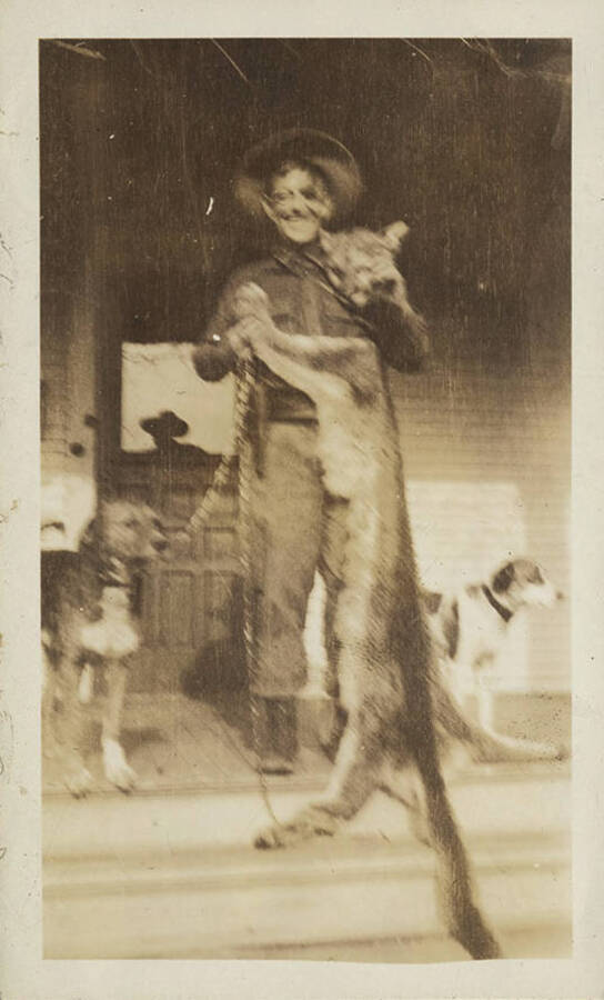 A hunter stands proudly with a harvested cougar on the front porch. Hunting dogs stand nearby.