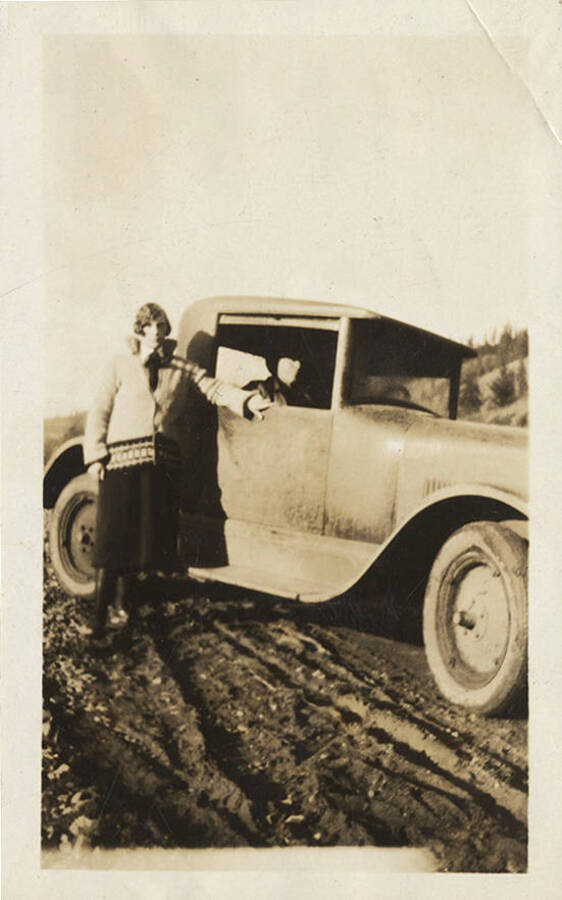 A woman poses next to an automobile on a dirt road.