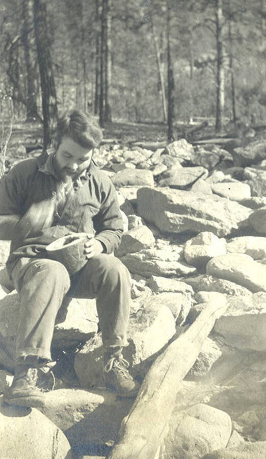 A young man sits on a pile of rocks and eats from a bowl.