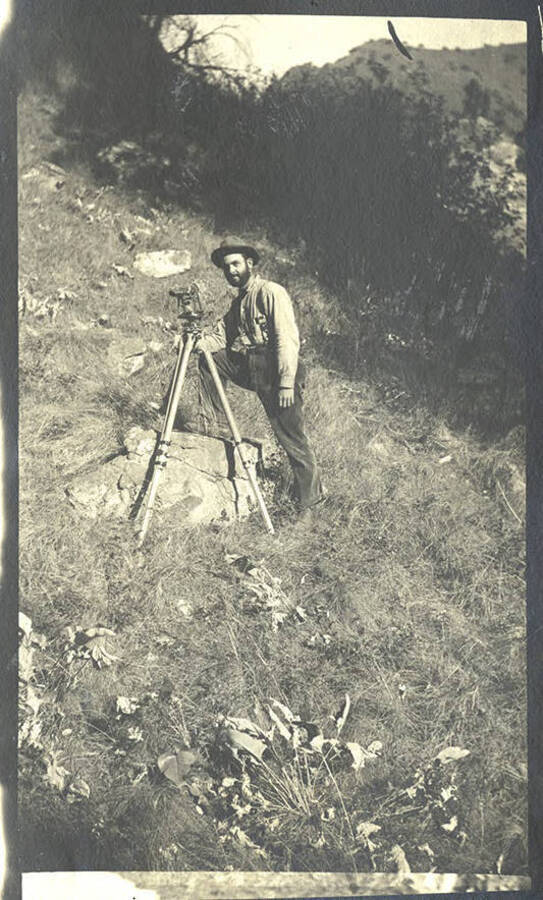 A man stands behind surveying equipment on a hill.