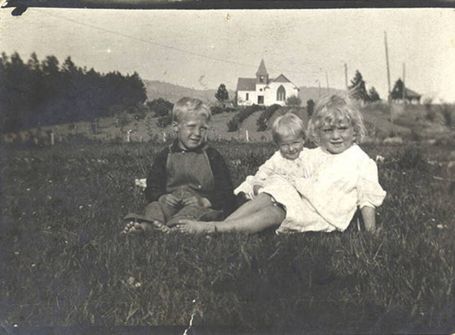Three children pose for a photograph in a grassy area with a white church in the background.