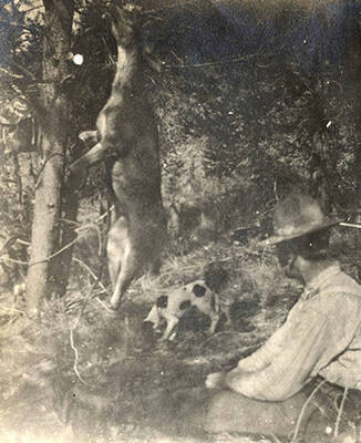 A man sits near a tree with a hanging deer ready for processing. A dog can be seen under the deer.