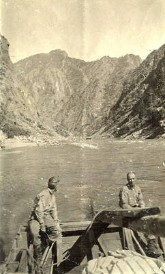 Two men sit on the back of a boat on the river with a view of the mountains in the background.