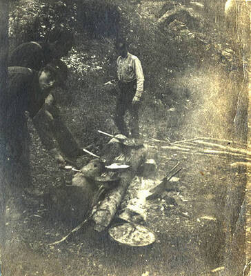Sumner Stonebraker (left) and two unidentified men cook over a campfire with frying pans. The photo caption reads: 'One dozen.'