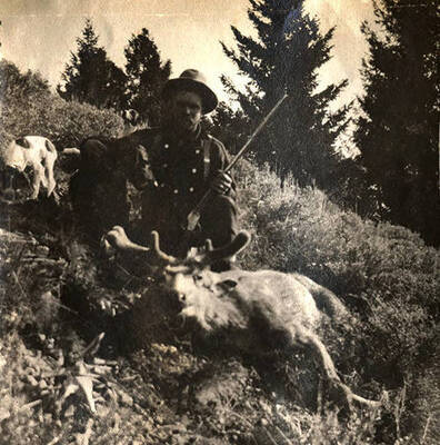 A man poses with a rifle and a harvested mule deer. Two dogs, including Juno, stand near the man.