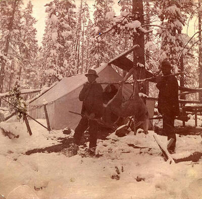 Two men pose with a harvester deer outside a canvas tent in the snow.