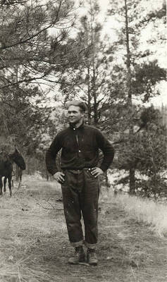 A hunter poses in a forested area. A horse can be seen in the background.