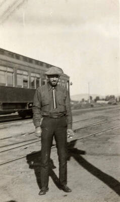 A man in a tie and a newsboy cap stands near the train tracks with a passenger car in the background.