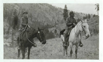 Two men ride horses and look around off into the distance.
