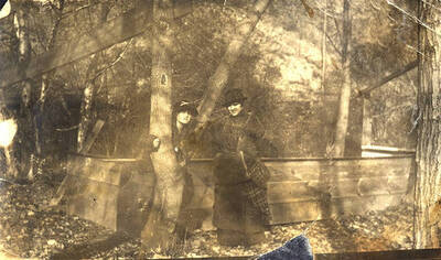 Two unidentified women pose near a wooden box structure among the trees in Stites, Idaho.