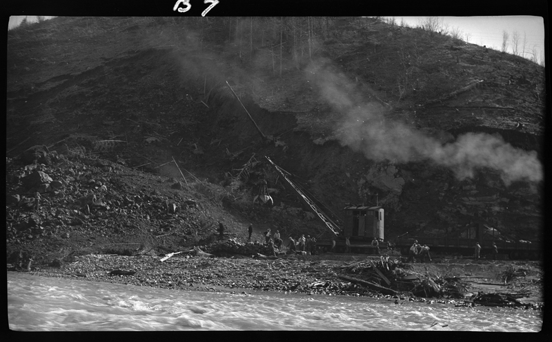 Negative of machinery at an unknown logging site. There is a crane-like piece of machinery in operation and workers surrounding it. There is a river between the photographer and the area that is being worked on.