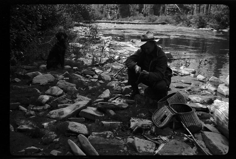 An unidentified man and a dog sit on the riverbank of the North Fork of the Coeur d'Alene River while the man is fishing. There is a fishing pole propped up in the rocks with the line cast into the water while the man works on something that isn't visible in his hands next to the fish baskets next to him.