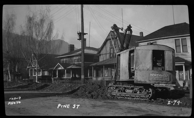 Photo of construction machinery on the side of the road of the 300 block of Pine Street in Wallace, Idaho. The machine reads "Northwest Engineering Co" on the side, and it appears to be working on the road. There is a line of houses on the opposite side of the road work.