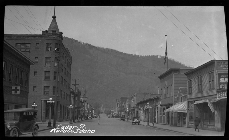 Photo of a Cedar Street looking West from near 7th Street in Wallace, Idaho taken circa 1925. People, cars, buildings, and an American flag are all visible, and beyond the town a tree covered mountain can be seen. 